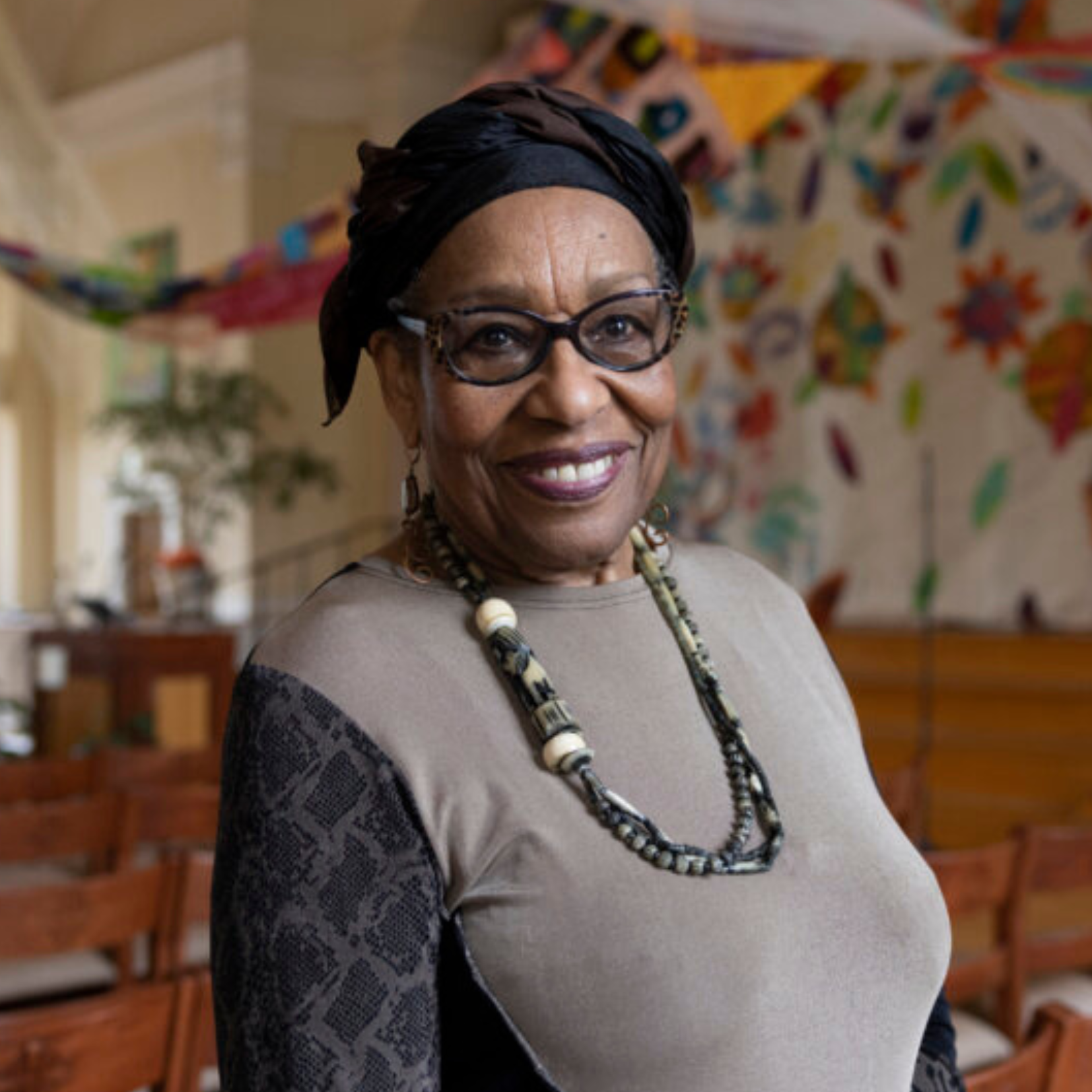 A smiling older woman with glasses, wearing a headscarf and a beaded necklace, stands in a room decorated with colorful hanging banners and wooden chairs, featured in our Donor Spotlight honoring the Walter L. Walker Legacy Fund.