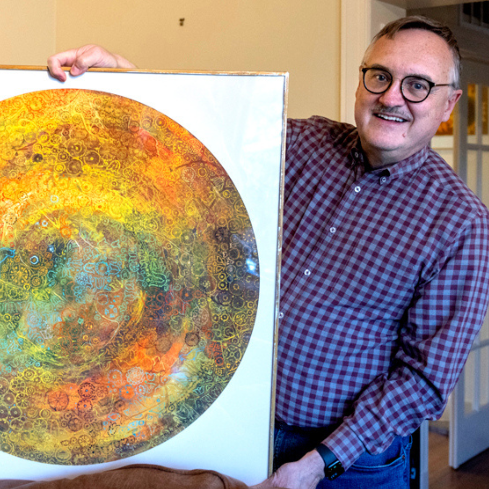 A smiling man in glasses and a plaid shirt holds up a colorful abstract circular artwork with swirling patterns—his Visionary Bequest—while standing indoors near a door.