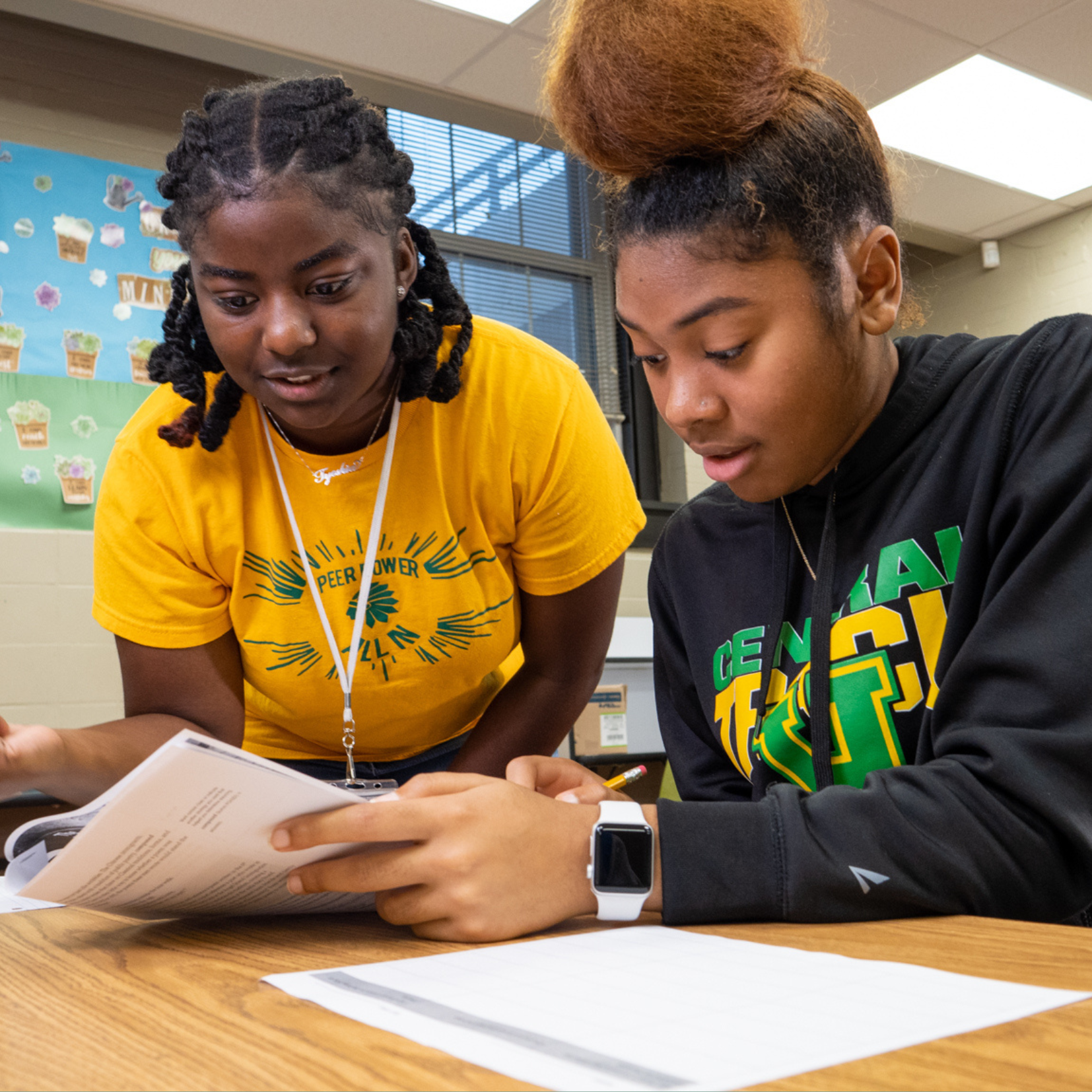 Two Memphis students sit at a desk in a classroom, looking at an open book together. One wears a yellow shirt and points at the book, while the other, in a black sweatshirt, listens and holds a pencil over lined paper.
