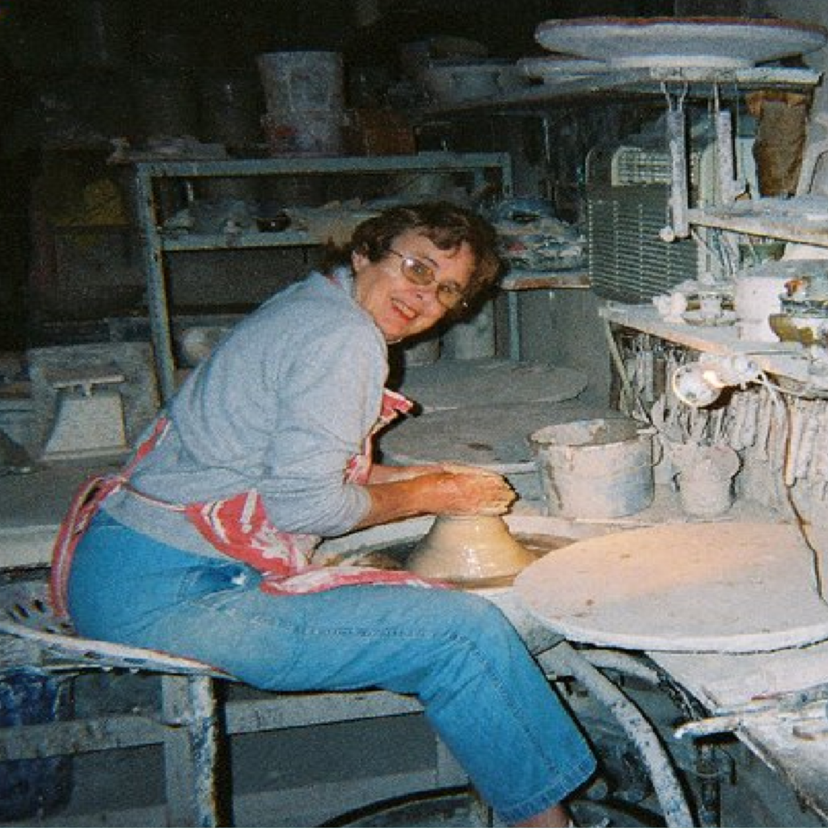 A person wearing glasses and an apron smiles while shaping clay on a pottery wheel in a Memphis workshop, embodying the spirit of Artists Supporting Artists amid shelves filled with pottery tools and ceramics.
