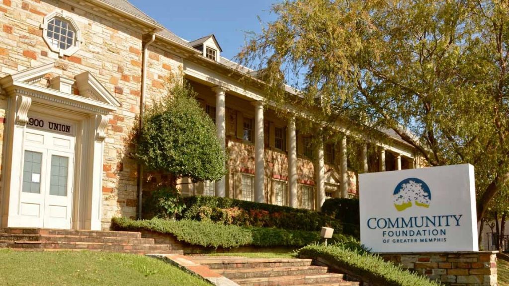 A stone building with columns and a sign in front that reads Community Foundation of Greater Memphis; trees and shrubs surround the entrance at 1900 Union, temporarily relocated from Crosstown Concourse.
