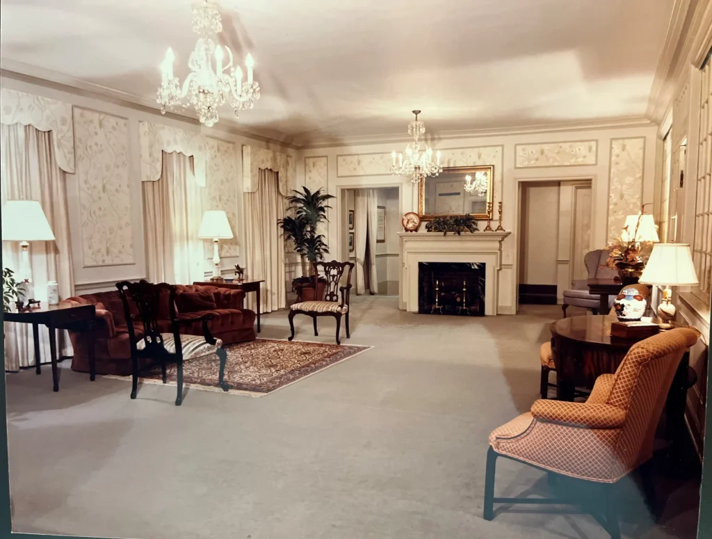 Elegant living room with chandeliers, ornate wall moldings, a fireplace with a large mirror above, upholstered chairs, a sofa, lamps, and small tables arranged on a carpeted floor—perfect for relaxing after exploring Crosstown Concourse.