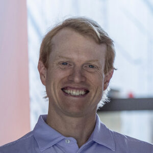 A man with light skin and short reddish-blond hair smiles broadly. He wears a lavender collared shirt and stands in front of a softly blurred indoor background.