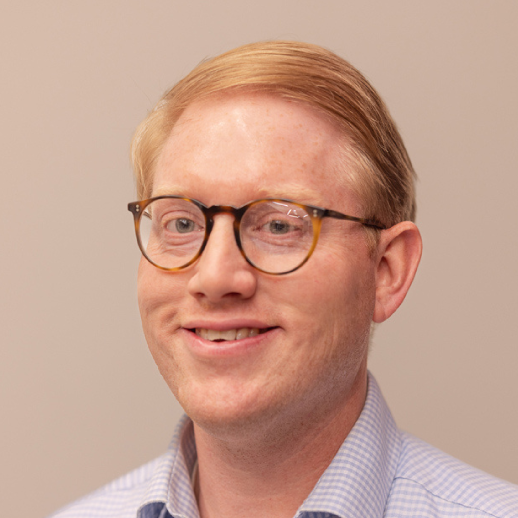 A man with short, light brown hair and glasses smiles at the camera. He is wearing a light blue, checkered button-up shirt against a plain beige background—Paul Shifflet shares insights on investing in Memphis.