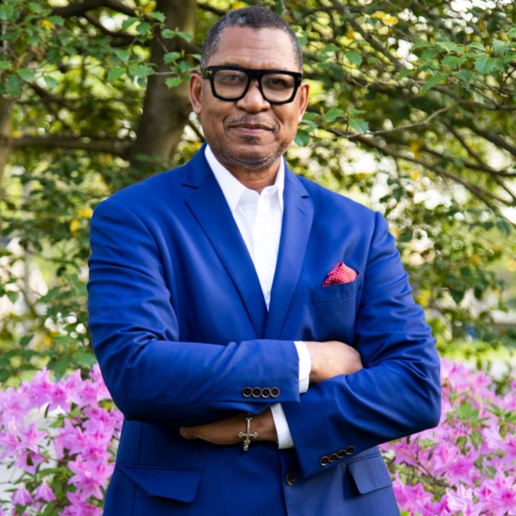 Antonio Tillis, in a blue suit with a red pocket square, stands outdoors with his arms crossed and smiling, reflecting his passion for giving back. Behind him are blooming pink flowers and green trees.