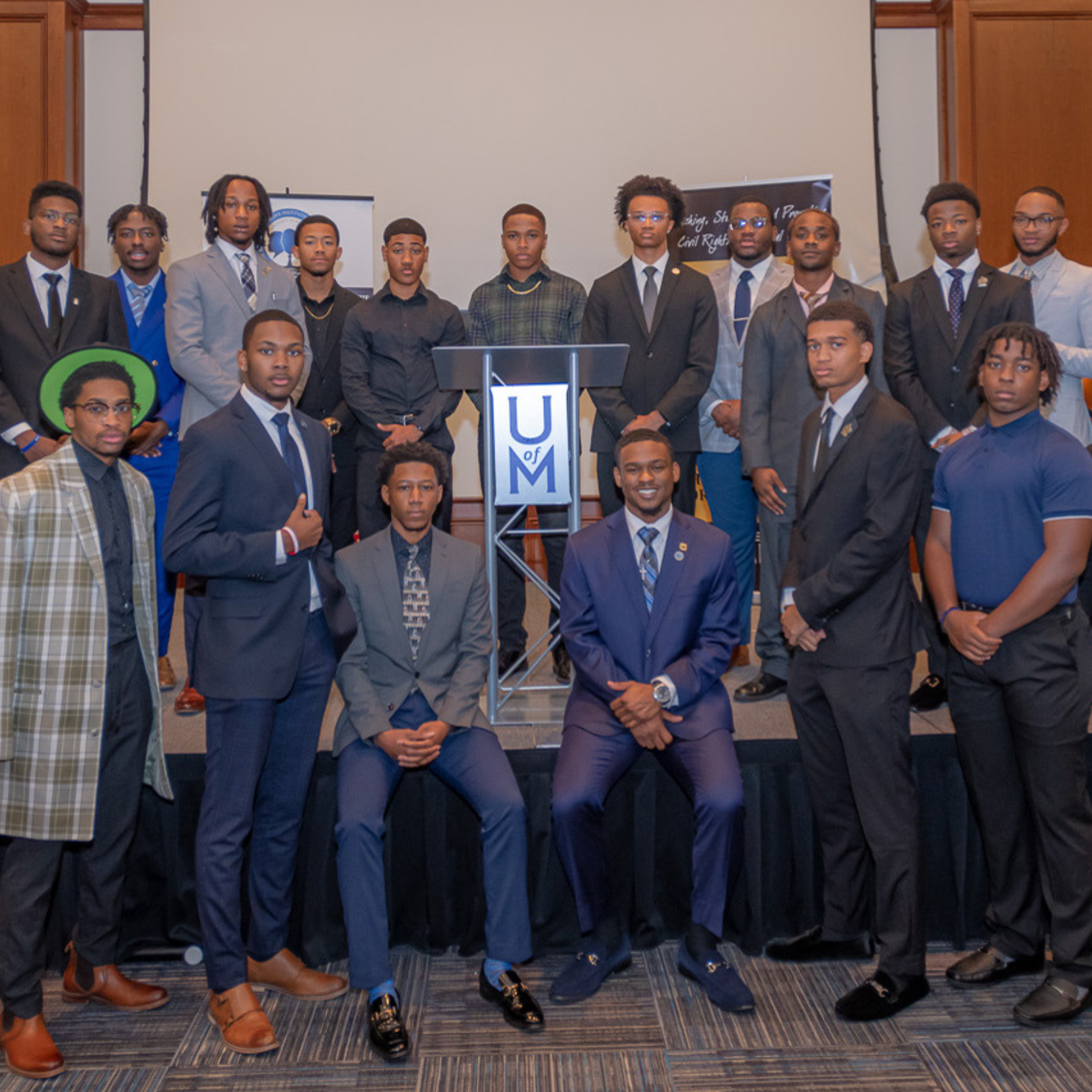 A group of 17 well-dressed young men pose in suits and formal attire around a podium with the UM logo, celebrating their achievements as HAAMI grant recipients, standing and sitting together on a stage in a formal setting.