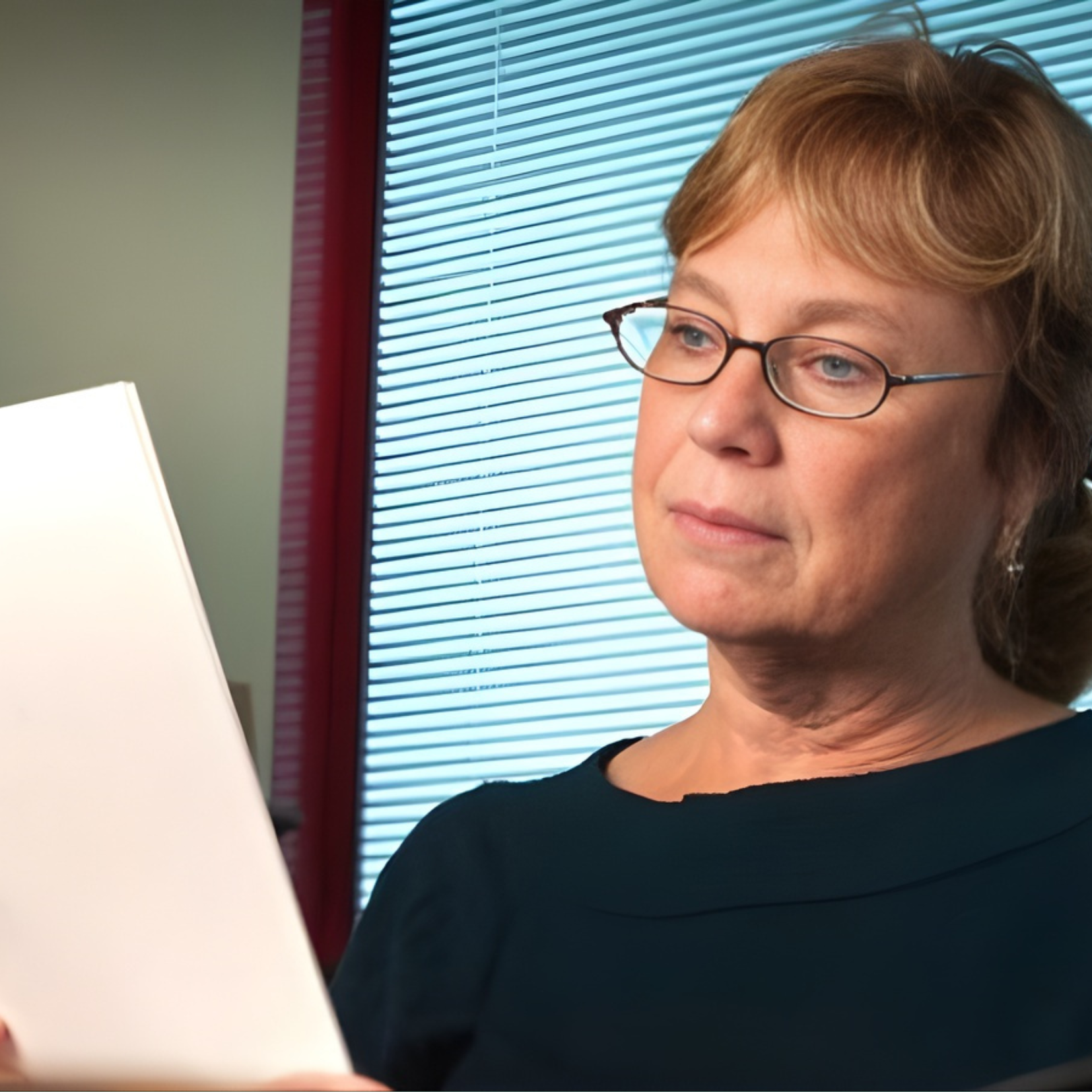 Linda Hendershot, on her philanthropic journey, sits indoors by a window with closed blinds. With short, light brown hair and glasses, she appears focused as she reads a document in her dark top.