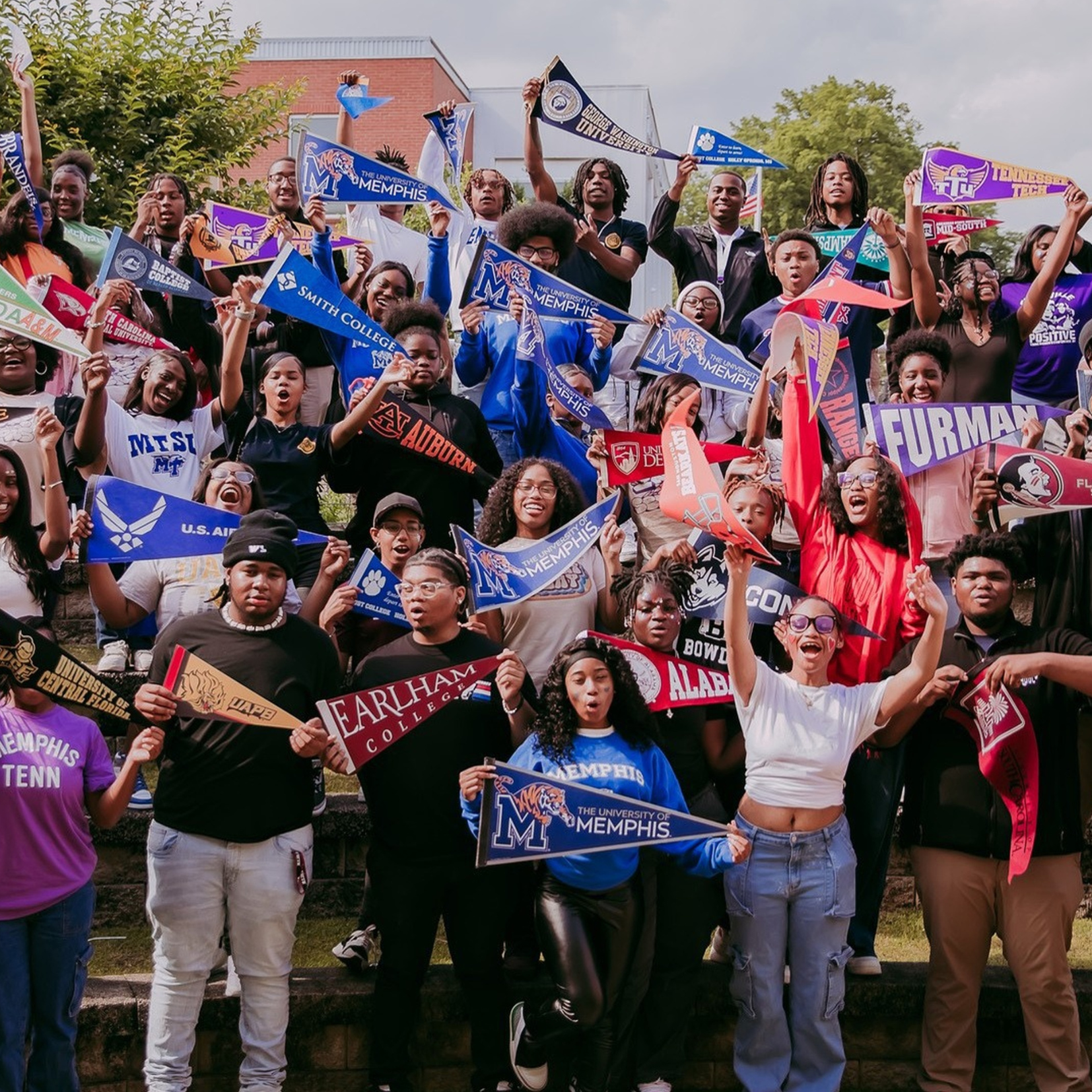 A large group of Soulsville students stands on steps outdoors, smiling and holding college banners, celebrating their future plans. Many wear shirts representing different colleges—shaping futures with strategic support. Trees and a building are in the background.