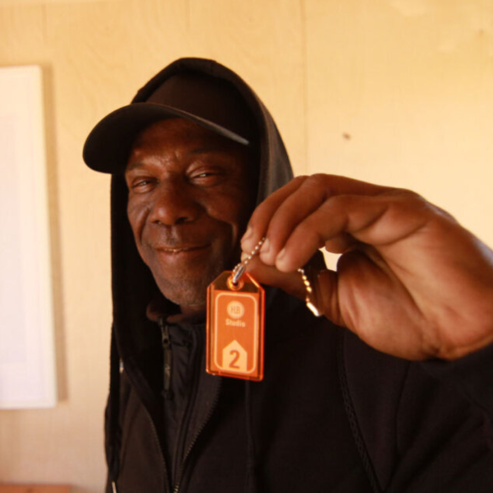 A person wearing a black hoodie and cap smiles while holding up a keychain labeled Studio 2 close to the camera, highlighting the importance of support services for vulnerable populations.