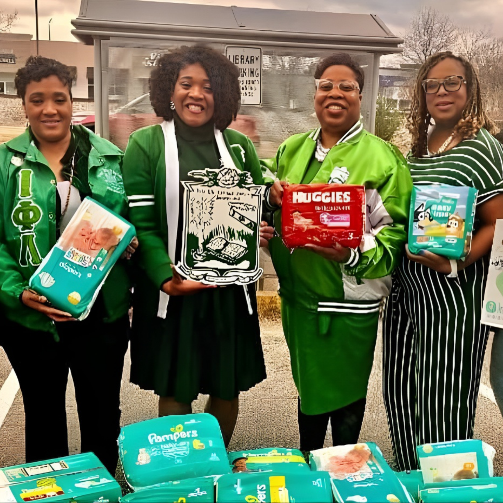Four women stand outdoors holding packages from Sweet Cheeks Diaper Ministry, with more supplies on a table in front of them. They are smiling and wearing green clothing, suggesting participation in a community service or diaper ministry event.