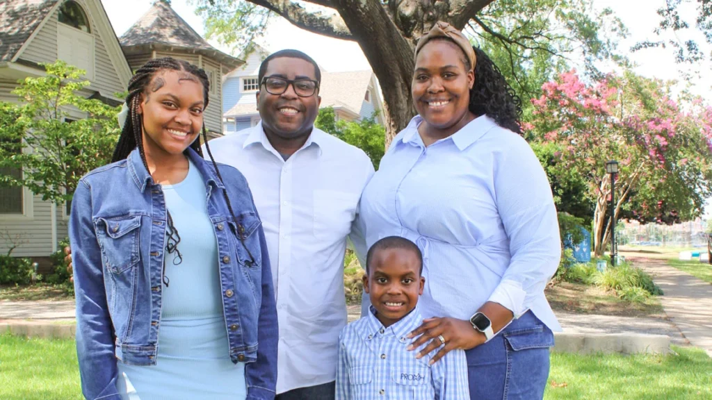 A smiling family of four poses outdoors in front of Hope House and a large tree, reflecting the warmth of community and care. The group includes two adults and two children, all wearing blue and white clothing on a sunny day.