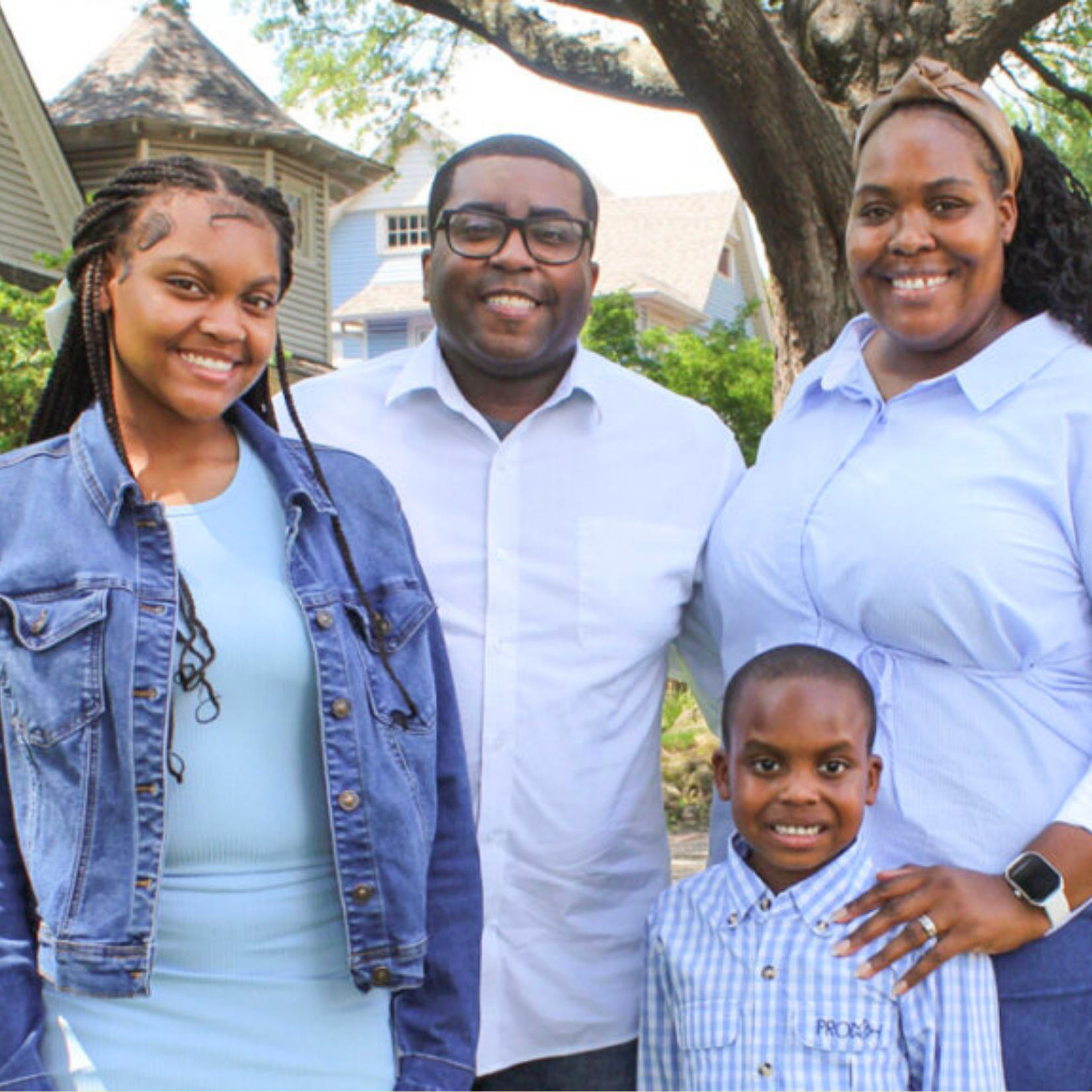 A smiling family of four poses outdoors, embodying the spirit of care and community. The parents stand behind, while their daughter in a denim jacket and young son in a blue-checkered shirt are in front, with trees and houses surrounding them.