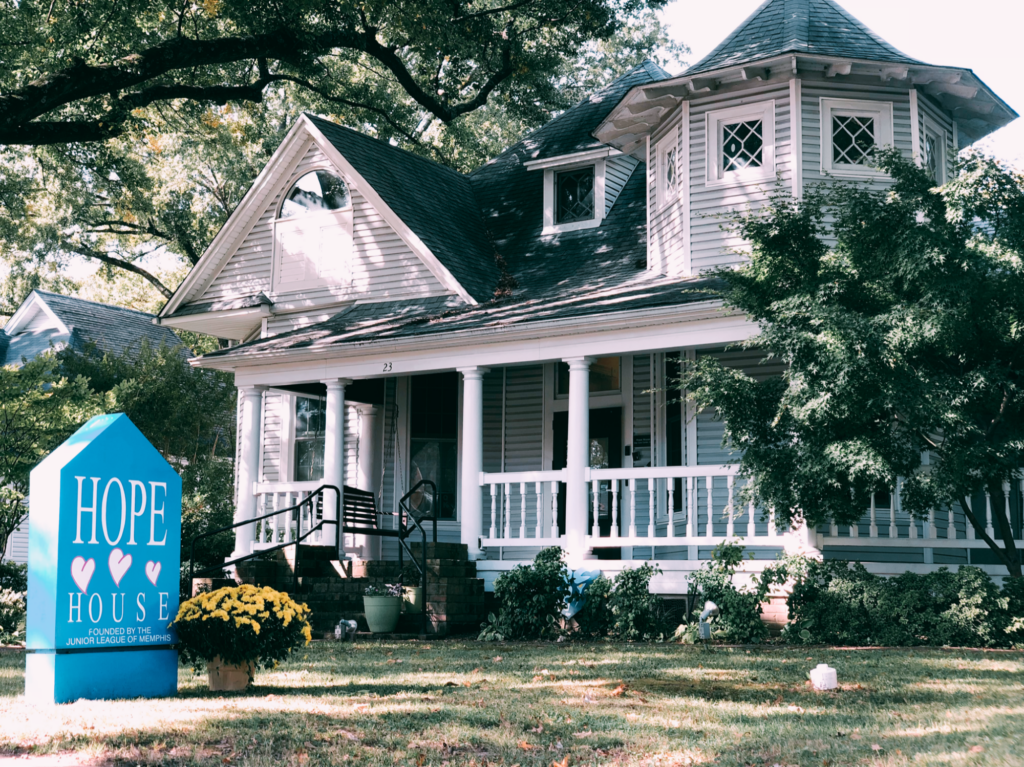 A large gray house with a wraparound porch and turret sits behind a blue Hope House sign with pink hearts, welcoming the community. The yard has green grass, a small tree, and potted yellow flowers by the porch steps.