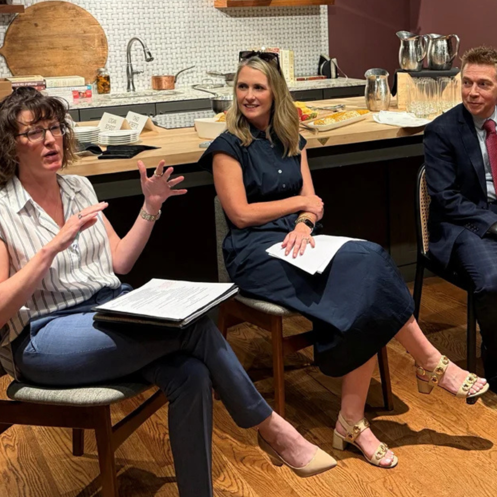 Three people sit on chairs in a kitchen-like setting, engaged in conversation about activating their personal giving plan. One woman gestures as she speaks, while the others listen attentively, holding papers. Drinks and snacks are visible on the counter behind them.