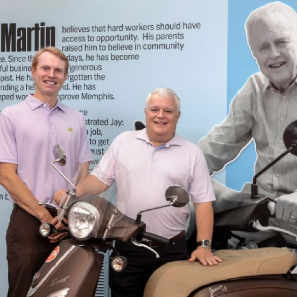 Two smiling men stand next to each other behind scooters. Behind them, a large wall displays text and a black-and-white photo of an older man smiling in a button-up shirt, celebrating the Martin family legacy and generational giving.