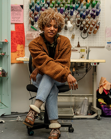 A person with curly hair sits crossed-legged on a chair in a sewing studio, surrounded by colorful thread spools and fabric, wearing a brown jacket, jeans, and boots.