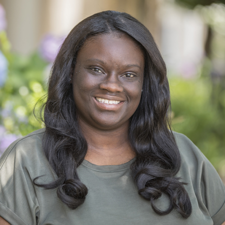 Aerial Ozuzu, with long, wavy dark hair, smiles while standing outdoors in a green t-shirt. Greenery and blurred flowers provide a vibrant background.