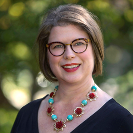 Aerial Ozuzu, a woman with short brown hair and glasses, smiles outdoors. She wears a black top and a colorful necklace with turquoise and red beads, set against a backdrop of blurred greenery.