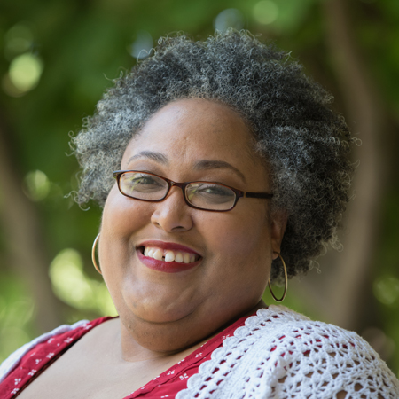Aerial Ozuzu, a woman with short curly gray hair, wearing glasses, gold hoop earrings, a red top with white patterns, and a white crocheted shawl, smiles outdoors against a softly blurred green background.