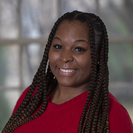Aerial Ozuzu, with long, braided hair, smiles at the camera. She is wearing a red sweater and stands in front of a window with a softly blurred outdoor background.