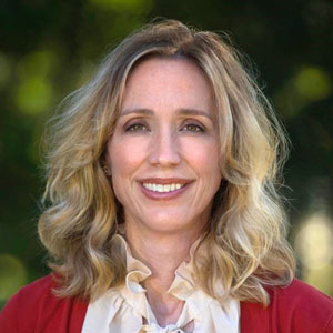 Aerial Ozuzu, a woman with wavy blonde hair, wearing a cream blouse and a red blazer, smiles in an outdoor setting with blurred greenery in the background.