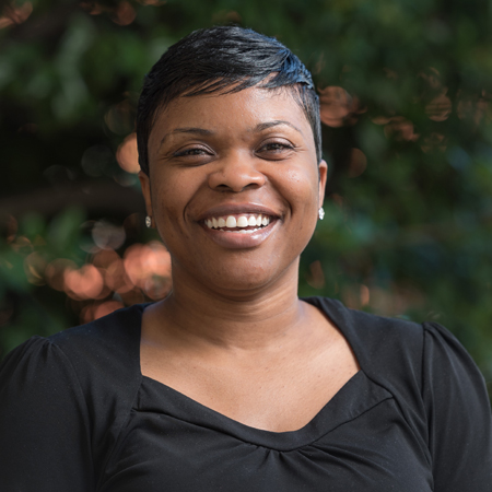 Aerial Ozuzu, a woman with short black hair and wearing a black top, smiles warmly at the camera. The background is blurred with green foliage and soft lighting.