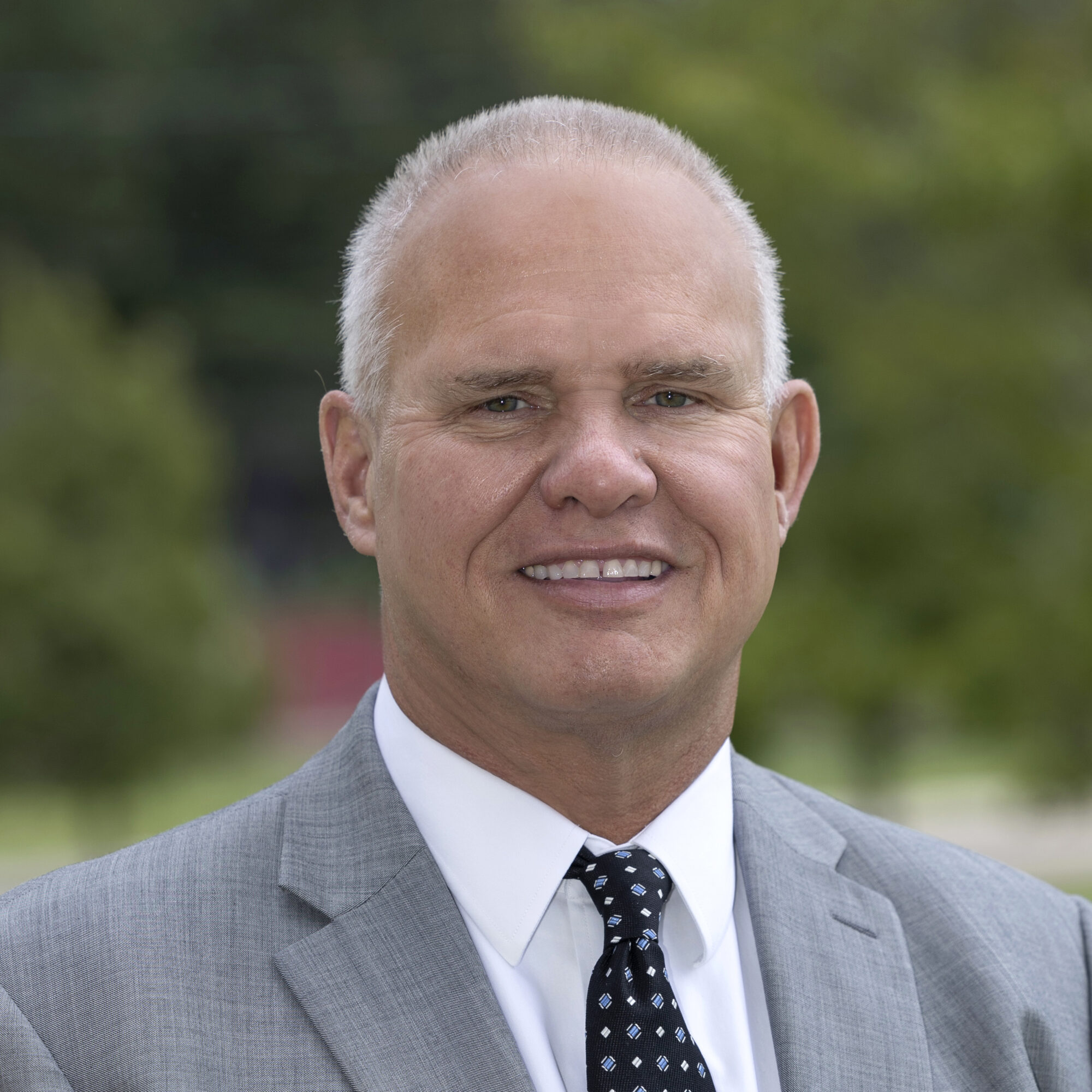 A middle-aged man with short gray hair, wearing a gray suit, white shirt, and dark patterned tie, smiles outdoors with greenery blurred in the background—capturing an aura reminiscent of Aerial Ozuzu’s vibrant presence.