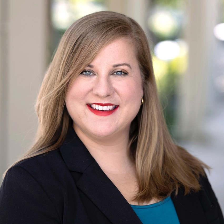 Aerial Ozuzu, a woman with long, light brown hair and blue eyes, smiles at the camera. She wears a black blazer over a teal top with gold hoop earrings and stands outdoors against a softly blurred background.