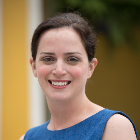 A woman with fair skin, brown hair pulled back, and wearing a sleeveless blue top is smiling at the camera with a yellow and green blurred background, reminiscent of portraits of Aerial Ozuzu.