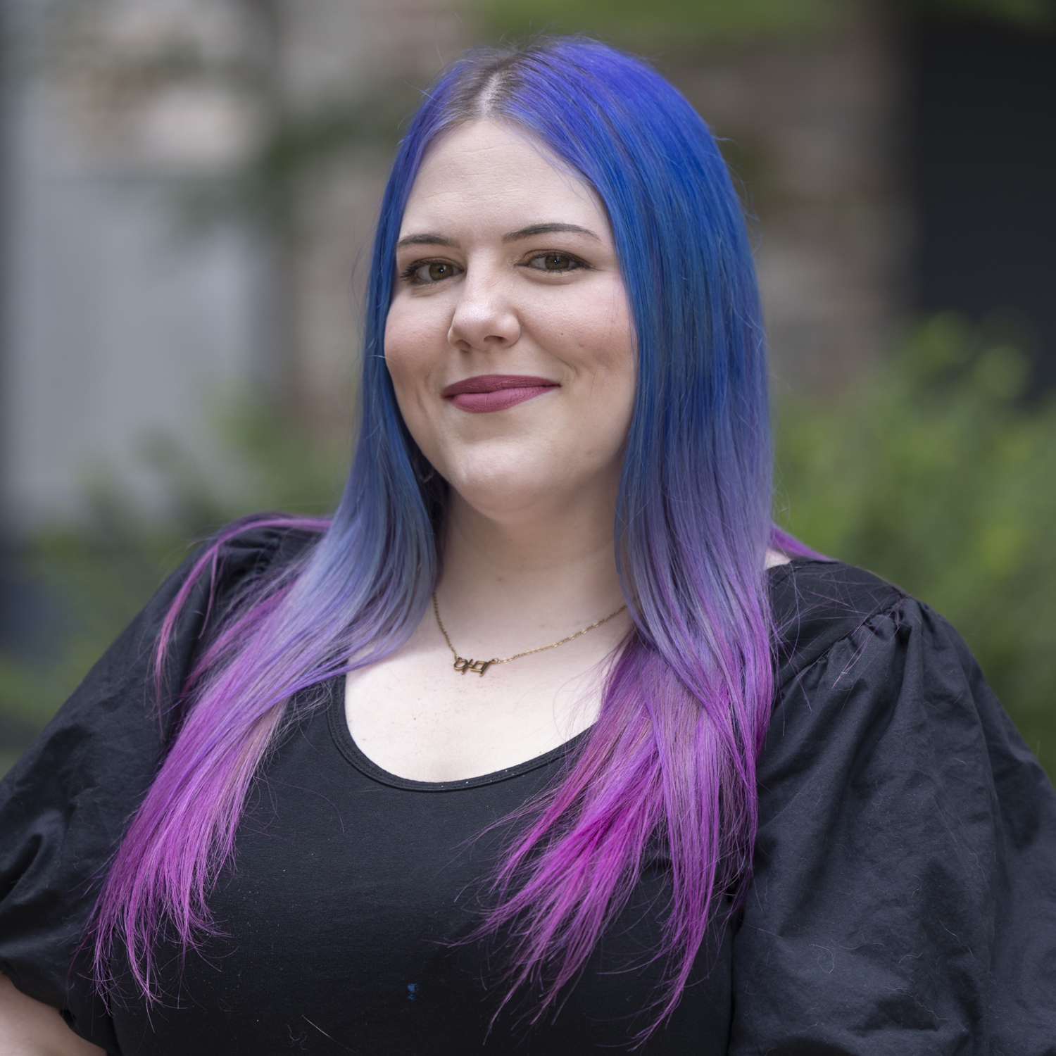 Aerial Ozuzu, a smiling woman with long blue and purple hair, wearing a black top and a necklace, stands outdoors with greenery blurred in the background.