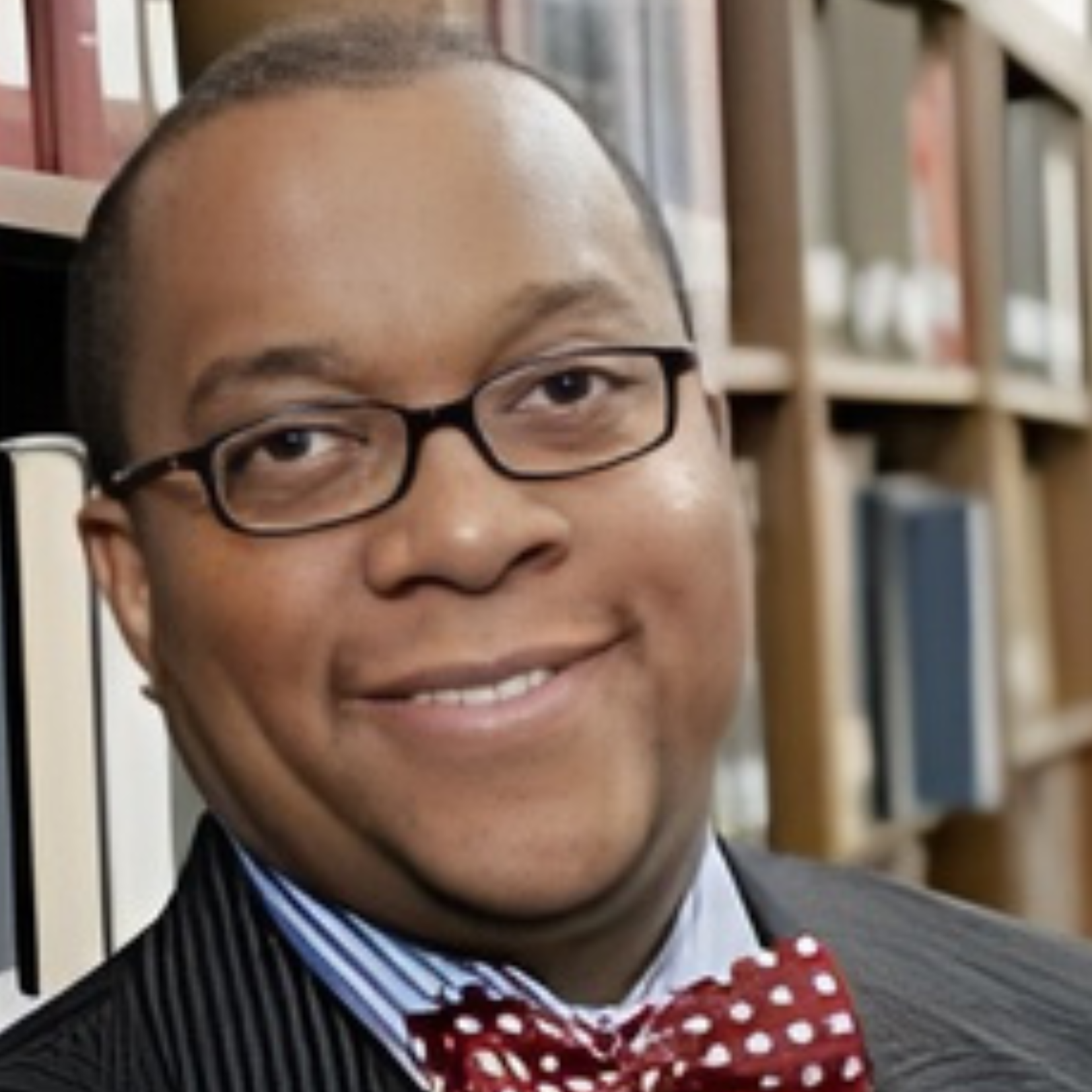 Charles McKinney, wearing glasses, a red polka dot bow tie, and a striped suit, smiles while standing in front of bookshelves in a library.