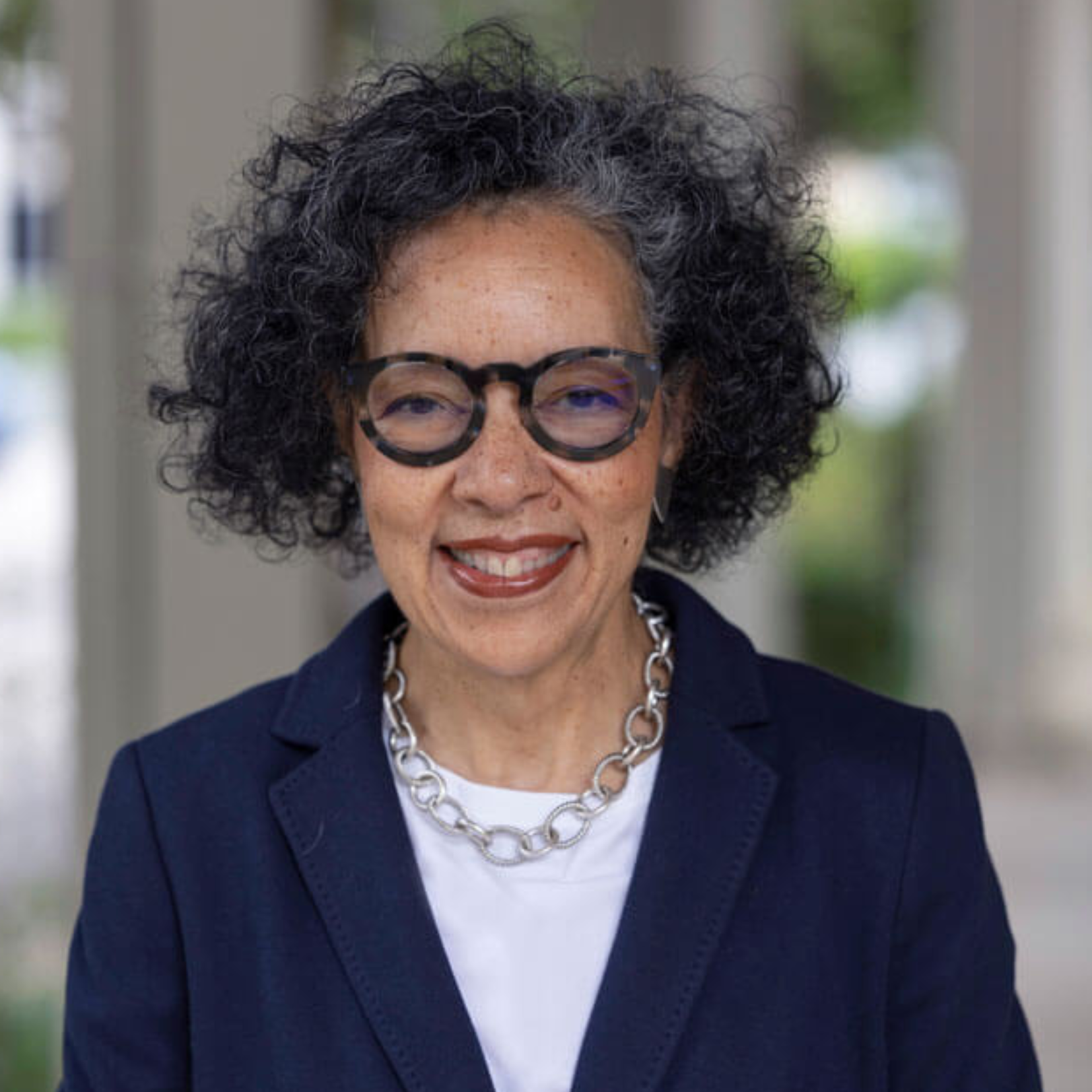 Yvonne Madlock, a woman with curly black and gray hair, wearing large round glasses, a navy blazer, a white top, and a chunky silver necklace, smiles while standing outdoors in a blurred setting.