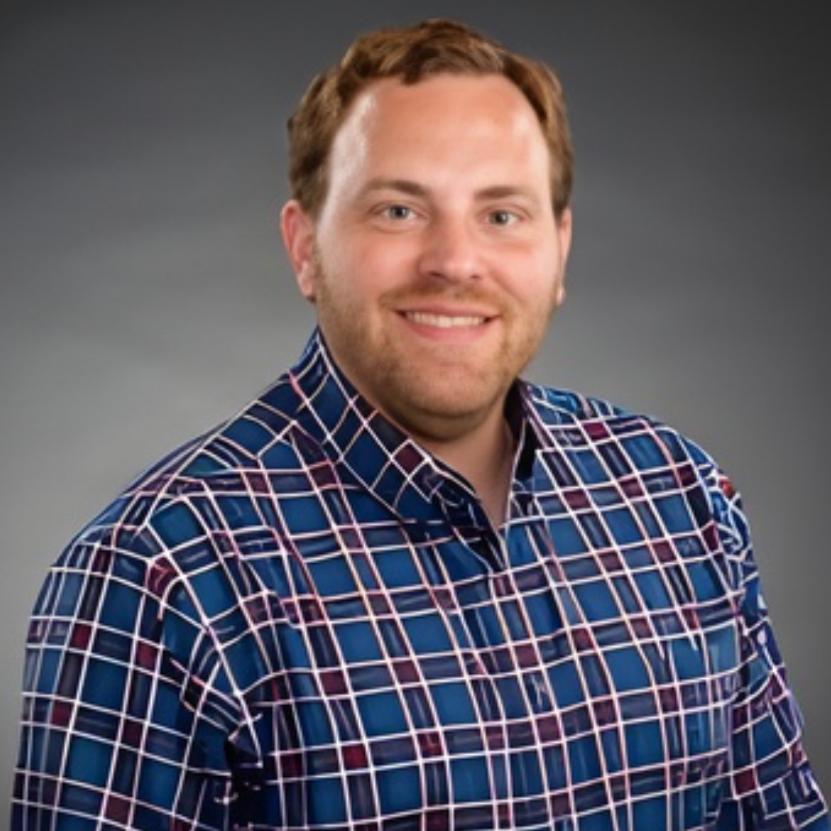 Jason Dapper, a man with short brown hair and a beard, smiles at the camera. He is wearing a blue shirt with a white and red plaid pattern, set against a plain gradient gray background.