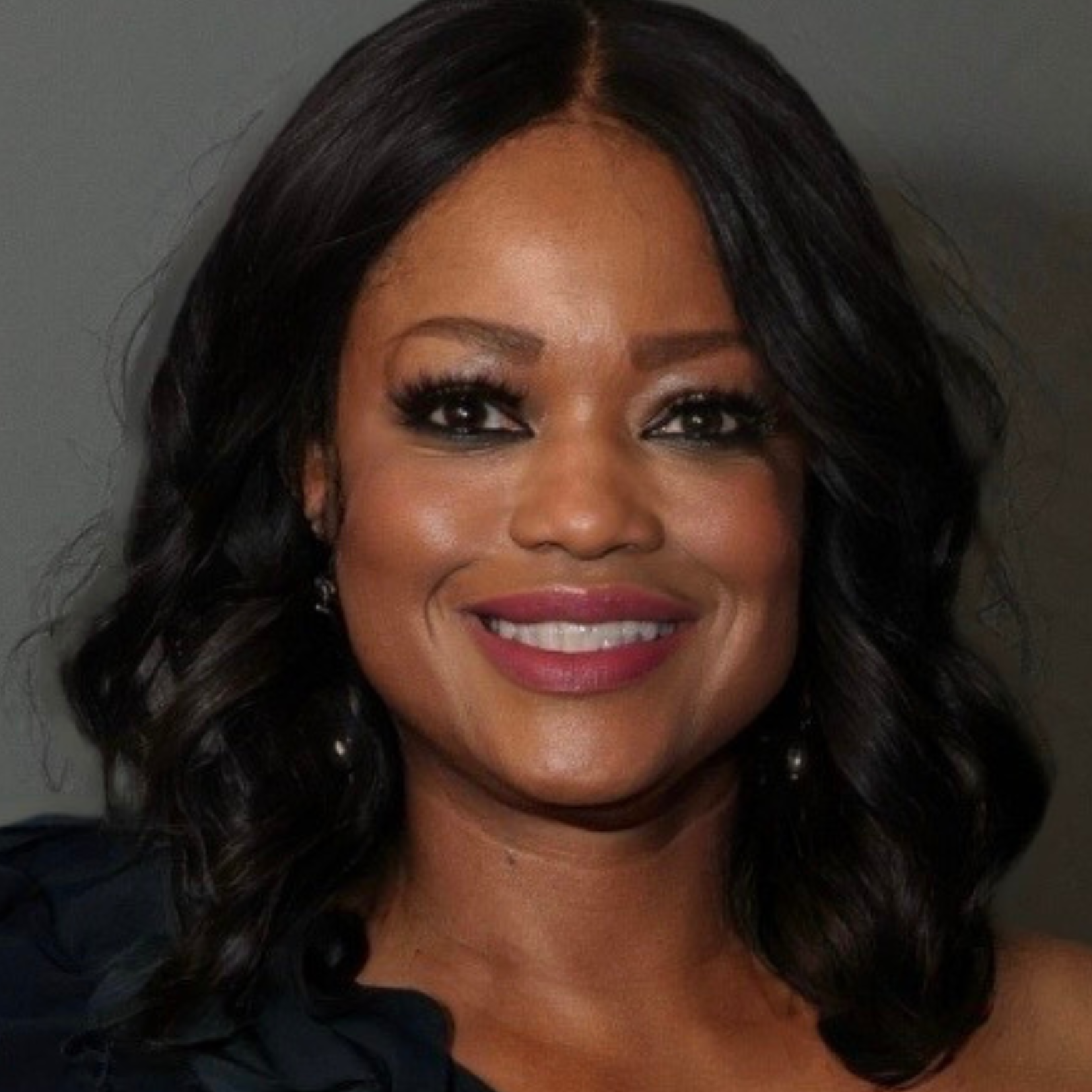 A woman with shoulder-length wavy black hair and glowing skin smiles warmly. She is wearing a dark, off-the-shoulder top and has subtle, elegant makeup with pink lipstick and prominent eyelashes.