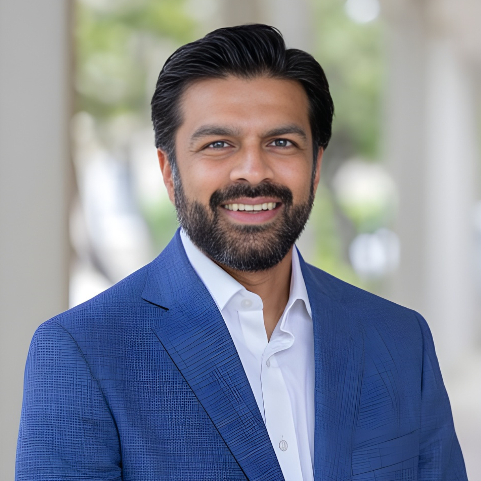 A man with dark hair and a beard, wearing a blue suit jacket and white shirt, smiles while standing outdoors with blurred greenery and columns in the background.