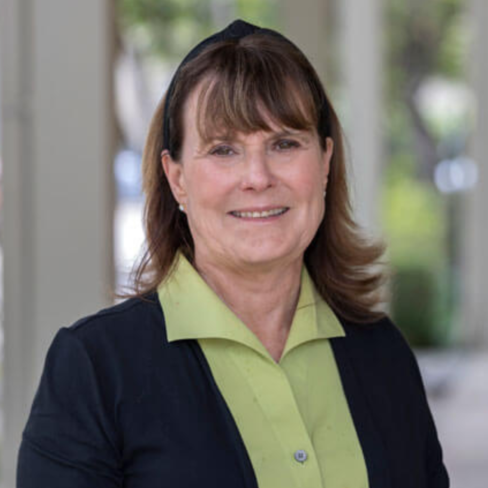 A woman with brown hair and bangs, wearing a light green blouse and black sweater, smiles while standing outdoors in a shaded walkway with blurred greenery in the background.