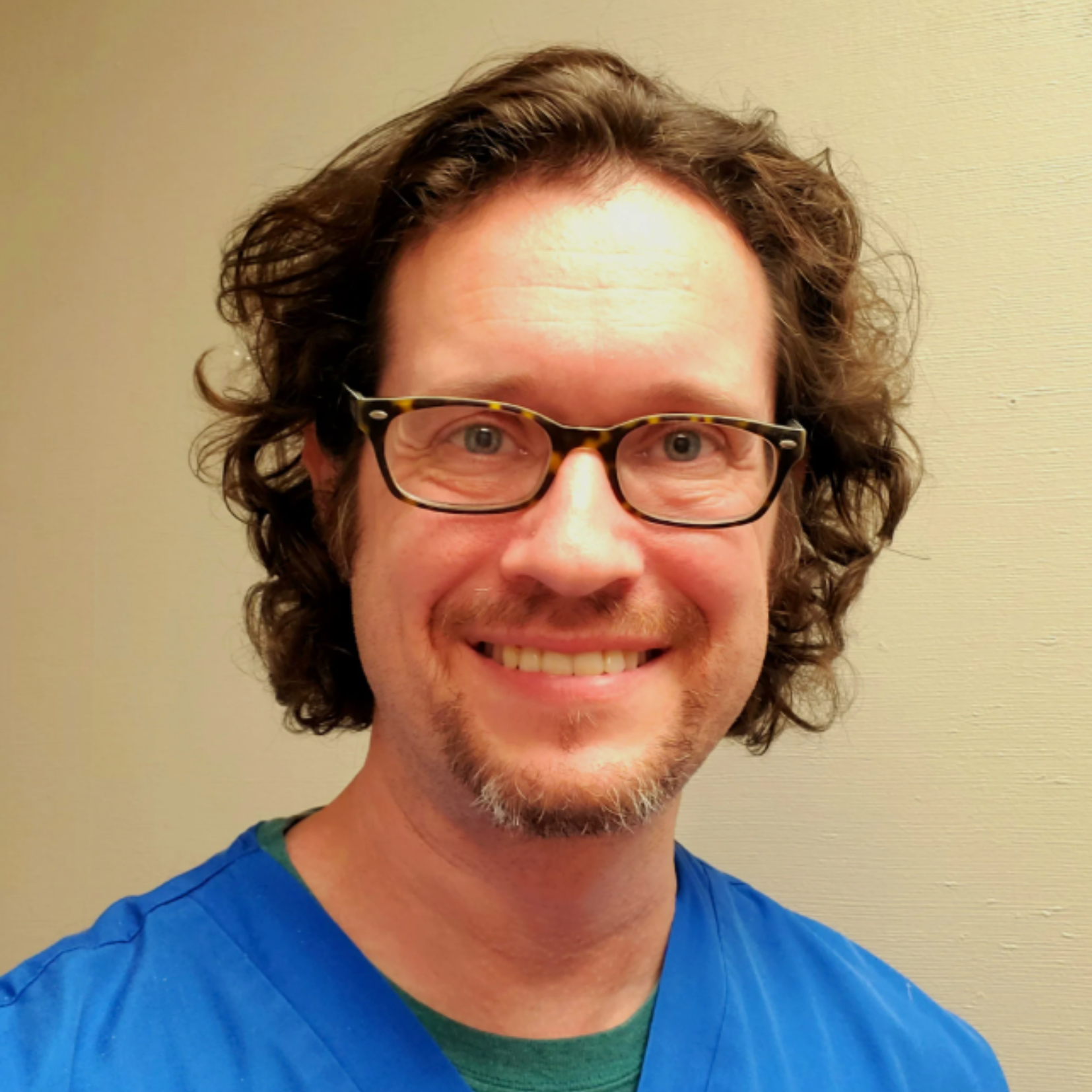 Louis Hamric, with curly brown hair, glasses, and a goatee, smiles at the camera while wearing blue medical scrubs. He stands confidently in front of a plain beige wall.