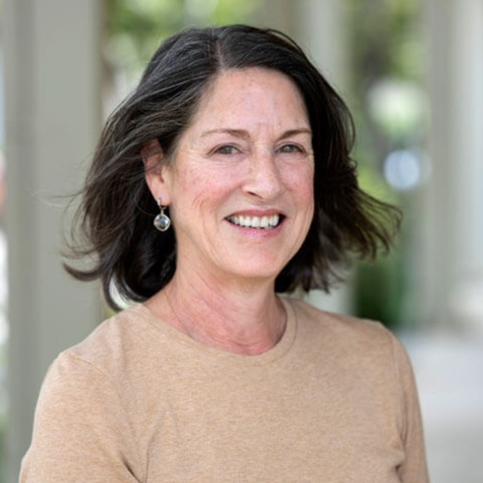 A middle-aged woman with shoulder-length brown hair is smiling at the camera. She is wearing a beige top and silver earrings, standing outside with blurred columns and greenery in the background.