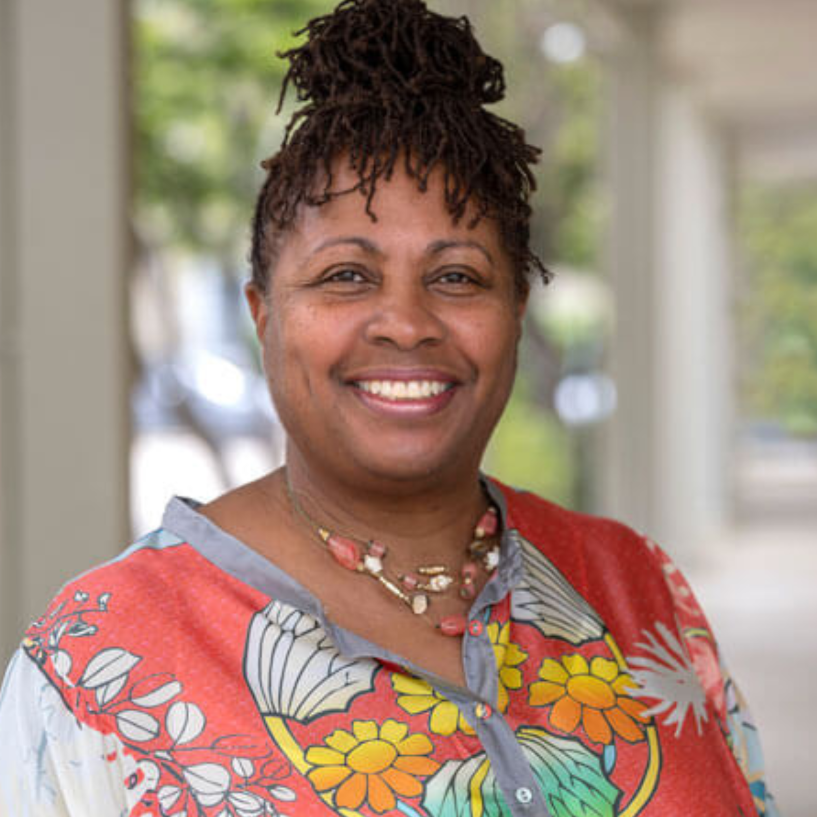 A woman with braided hair in an updo smiles outdoors, wearing a colorful blouse with flower patterns and a beaded necklace. The background is softly blurred with greenery and architectural elements, capturing Beverly Perkins' radiant presence.