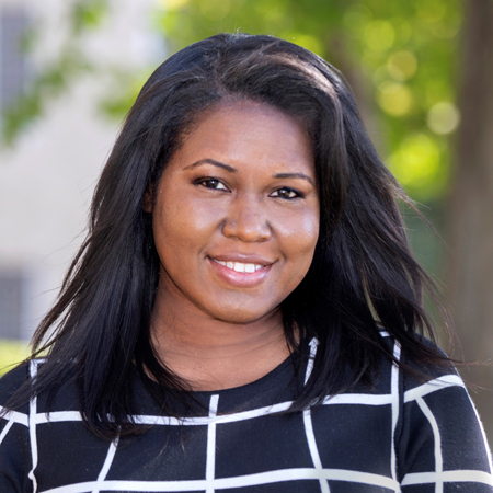 Aerial Ozuzu, a woman with long black hair, smiles outdoors in a black and white checkered top. Green trees and a building are blurred in the background.