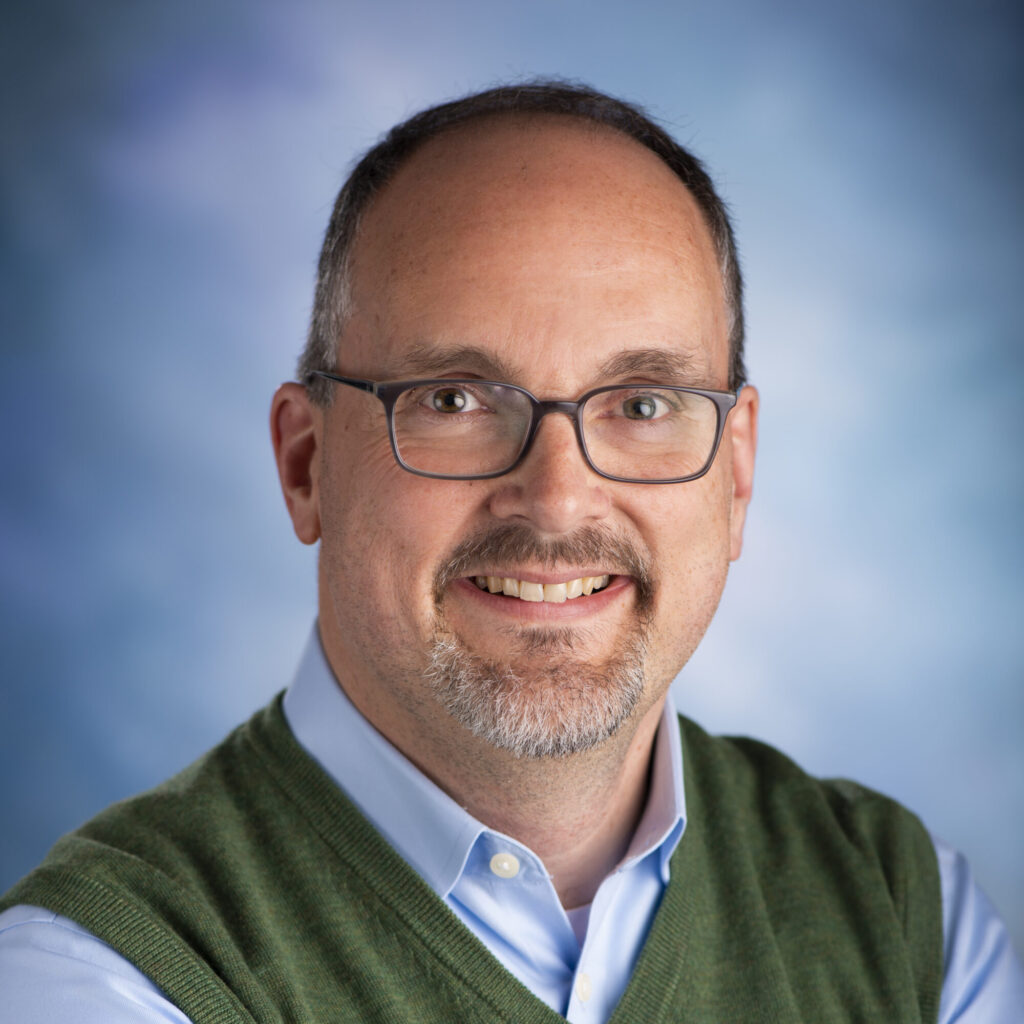A middle-aged man with glasses, short hair, and a goatee smiles at the camera. He wears a light blue shirt and green sweater vest against a blue background, representing Allied Professional Advisors at Seminar 2025.
