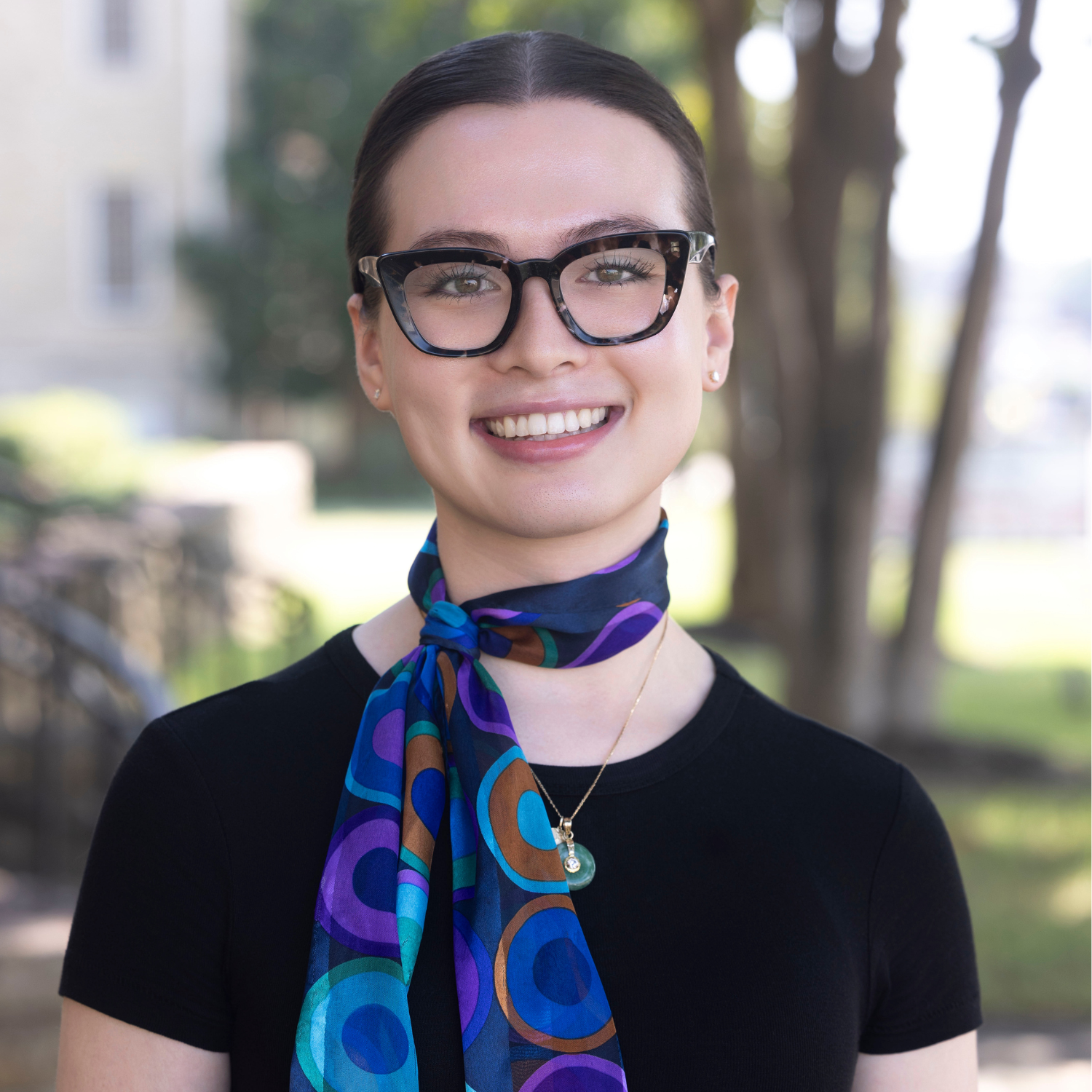 A person wearing glasses, a black top, and a colorful scarf smiles outdoors with trees and blurred buildings in the background, reminiscent of Randy Truong’s vibrant style.