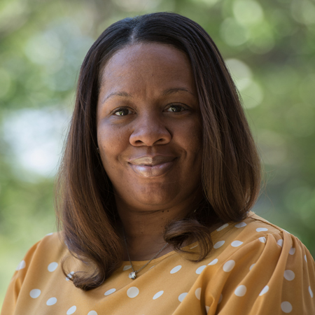 Aerial Ozuzu, with straight, shoulder-length hair, smiles gently in a yellow blouse with white polka dots. The blurred green and white bokeh background adds a soft touch.