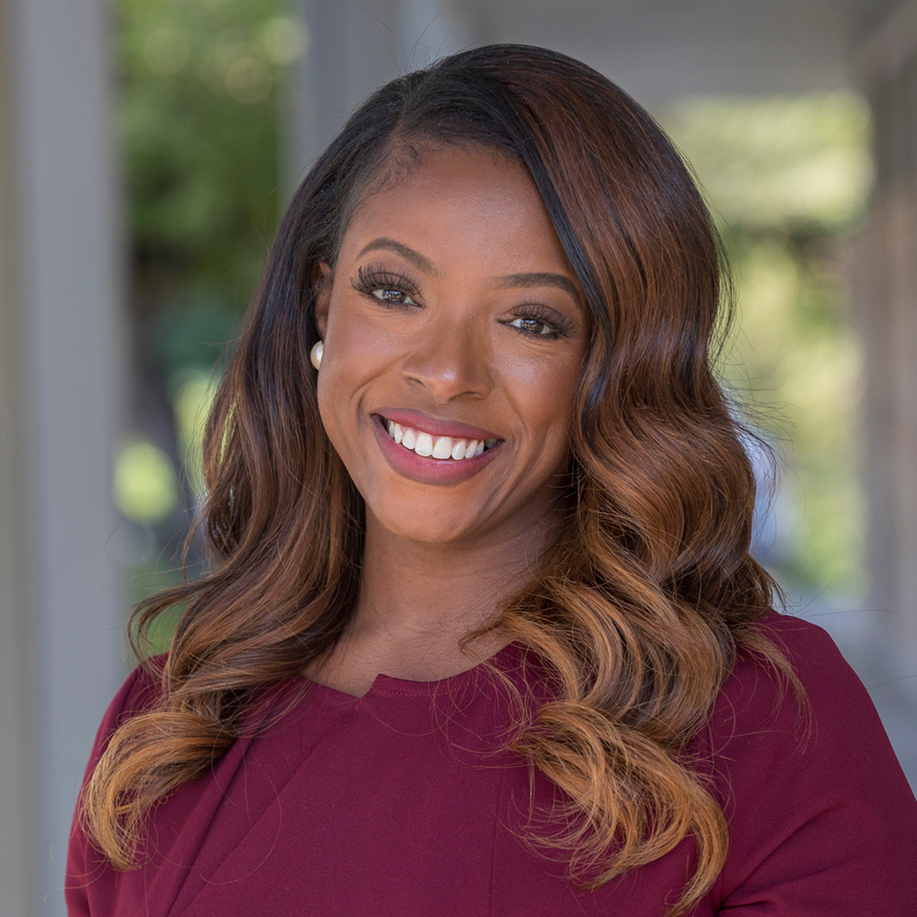 Aerial Ozuzu, with long, wavy brown hair and a maroon top, smiles outdoors. She wears pearl earrings and stands against a well-lit backdrop of greenery and columns.