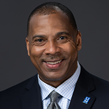 A smiling man, identified as Alex DeJarnett, in a dark suit, light blue checkered shirt, and gray tie poses against a dark background. He wears a pin with blue rectangles on his lapel.