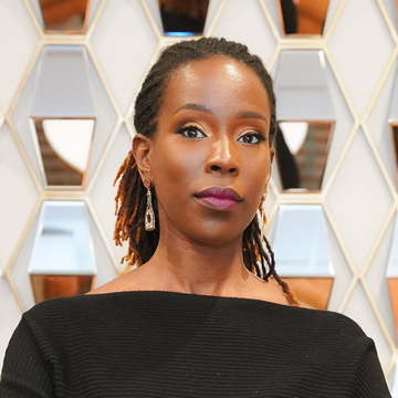 April Carter, with her long, styled hair, wears drop earrings and a black off-the-shoulder top as she stands before a geometric mirrored wall, looking confidently ahead with a neutral expression.
