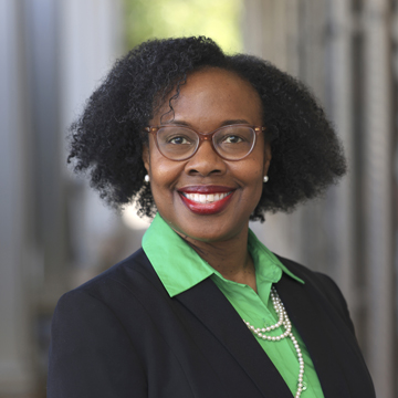 Belinda Simpson, with curly black hair, glasses, and red lipstick, smiles warmly. She wears a green collared shirt, black blazer, and pearl necklace. The softly blurred background hints at an outdoor hallway or corridor.