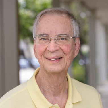 Bert Barnett, an elderly man with short gray hair and glasses, smiles warmly at the camera. He is wearing a light yellow polo shirt and standing outside, with greenery and columns blurred in the background.