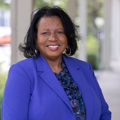 Carol Winfield Jones, a woman with dark hair, wearing a bright blue blazer and patterned blouse, smiles at the camera while standing outdoors in a shaded area.