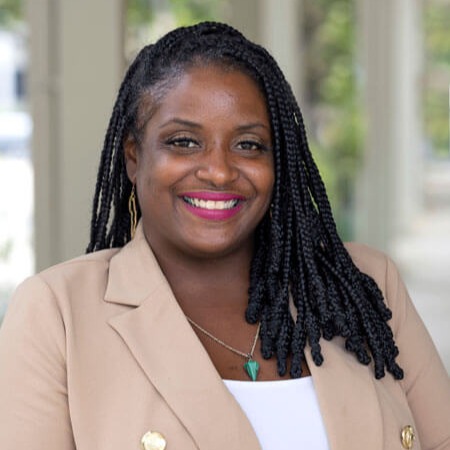 Crystal Chopin, a woman with long braided hair, smiles in a tan blazer over a white top, gold earrings, a turquoise pendant necklace, and bright pink lipstick, standing in a bright, blurred hallway.